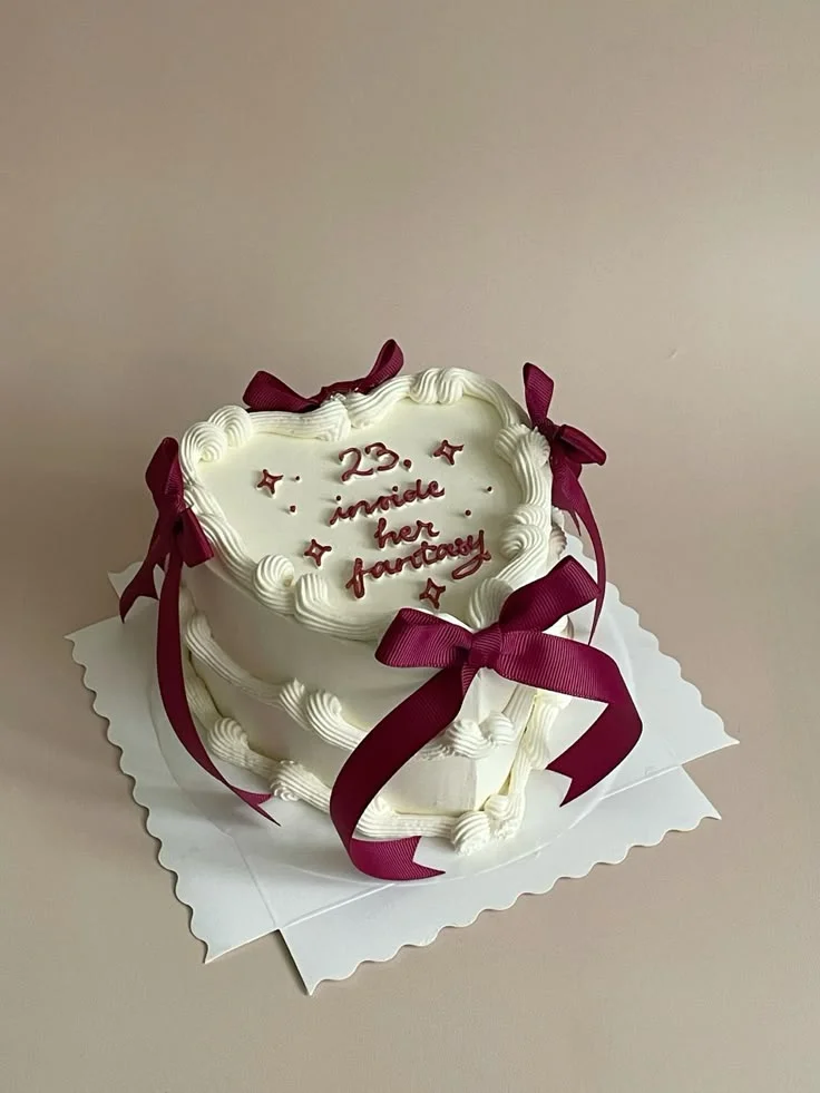 White heart-shaped minimalist cake with dark red piped icing text and matching satin ribbons on a white scalloped base. White heart-shaped minimalist cake with dark red piped icing text and matching satin ribbons on a white scalloped base.