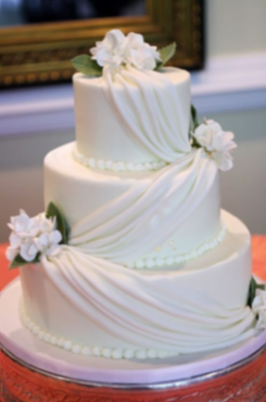 Elegant three-tier ivory fondant wedding cake featuring cascading white gumpaste fabric drapes and delicate white floral accents. Elegant three-tier ivory fondant wedding cake featuring cascading white gumpaste fabric drapes and delicate white floral accents.