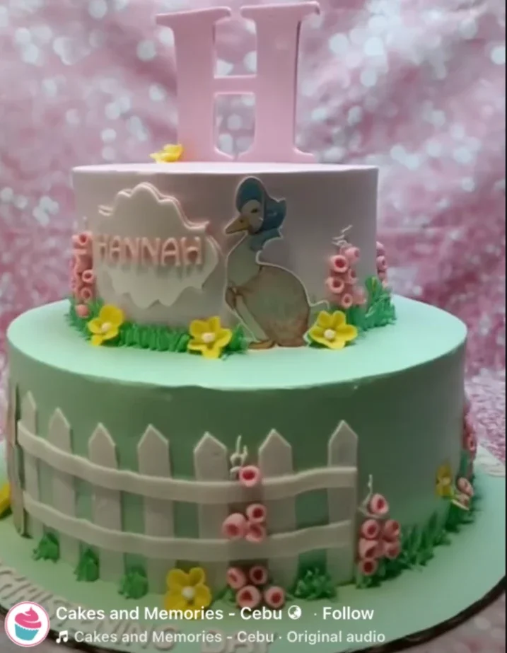 Two-tier Jemima Puddle-Duck themed cake in light pink and mint green with a large pink letter H topper and floral accents. Two-tier Jemima Puddle-Duck themed cake in light pink and mint green with a large pink letter H topper and floral accents.