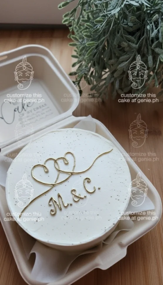 White minimalist bento cake with gold heart line art and gold initials M and C in a clamshell box. White minimalist bento cake with gold heart line art and gold initials M and C in a clamshell box.