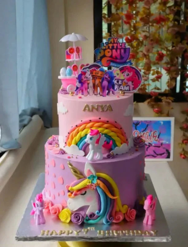 Two-tier My Little Pony themed cake in pink and purple with a large unicorn face, rainbow, and character toppers. Two-tier My Little Pony themed cake in pink and purple with a large unicorn face, rainbow, and character toppers.