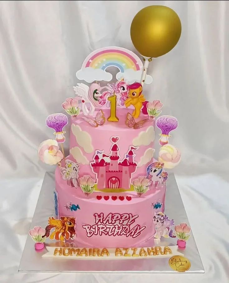 Pink 2-tier My Little Pony birthday cake with rainbow, castle, and character printouts, plus a gold balloon and number 1 candle. Pink 2-tier My Little Pony birthday cake with rainbow, castle, and character printouts, plus a gold balloon and number 1 candle.
