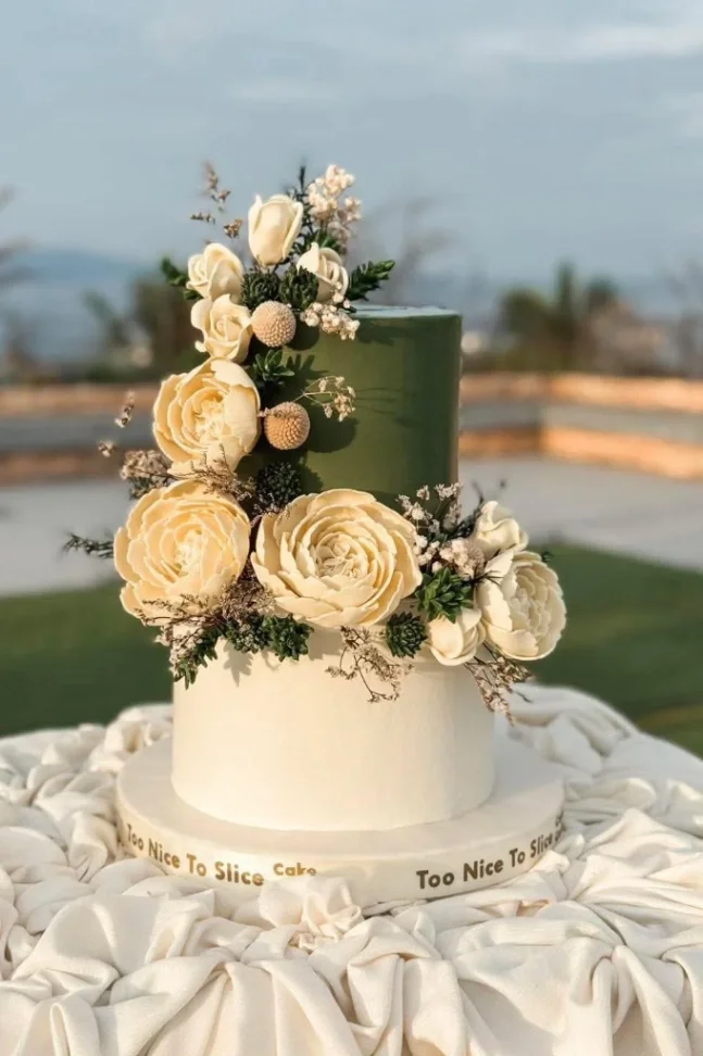 Elegant two-tier cake with olive green and ivory icing, decorated with large cream-colored gumpaste flowers and dried floral accents. Elegant two-tier cake with olive green and ivory icing, decorated with large cream-colored gumpaste flowers and dried floral accents.