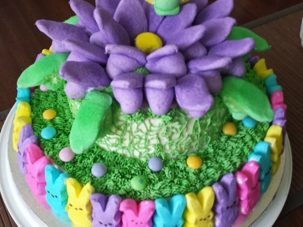 A colorful 1-tier cake decorated with purple marshmallow flower petals, green icing grass, and multi-colored Peeps marshmallow bunnies around the base. A colorful 1-tier cake decorated with purple marshmallow flower petals, green icing grass, and multi-colored Peeps marshmallow bunnies around the base.