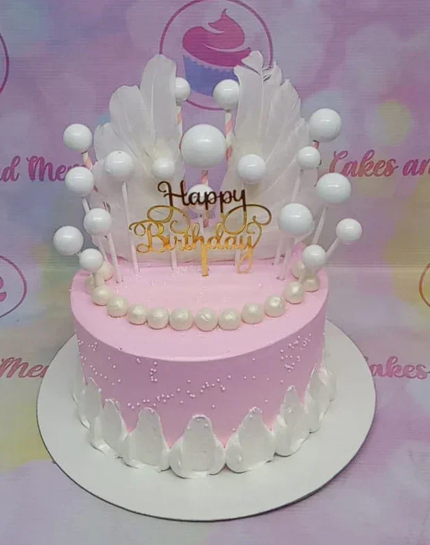 Pink birthday cake with white feather wings, gold Happy Birthday topper, and white decorative spheres. Pink birthday cake with white feather wings, gold Happy Birthday topper, and white decorative spheres.
