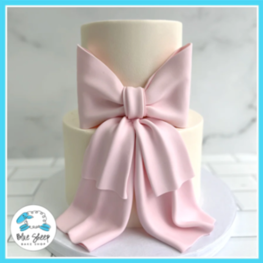 Elegant two-tier ivory fondant cake featuring a large, oversized pink fondant bow with long flowing ribbons draped down the front. Elegant two-tier ivory fondant cake featuring a large, oversized pink fondant bow with long flowing ribbons draped down the front.