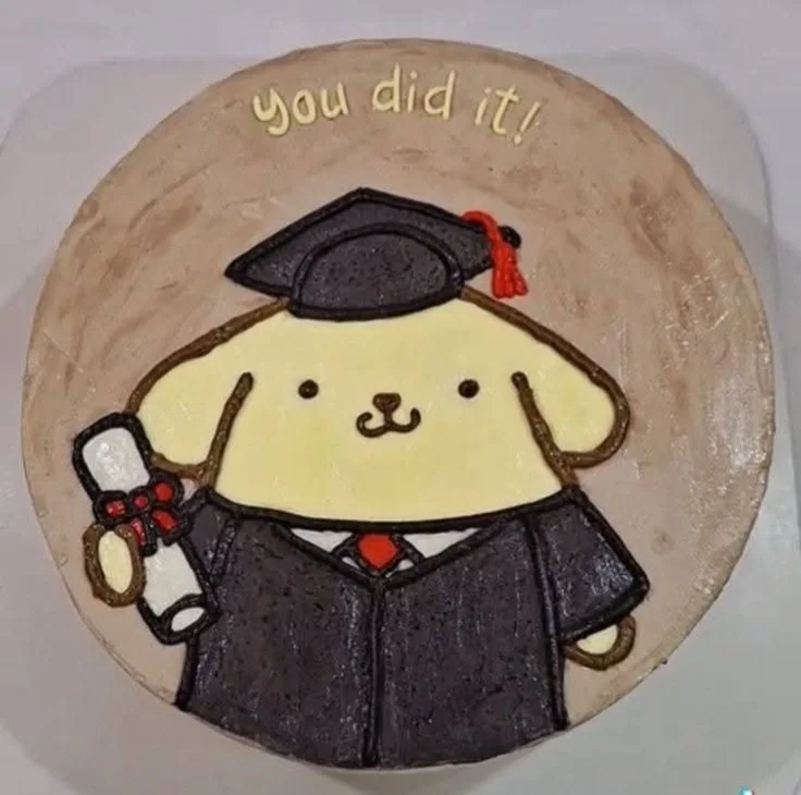 A round beige bento-style cake featuring a hand-piped Pompompurin character in a graduation cap and gown holding a diploma. A round beige bento-style cake featuring a hand-piped Pompompurin character in a graduation cap and gown holding a diploma.