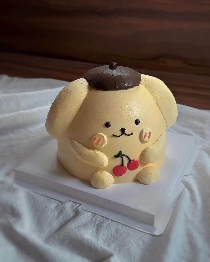 Pompompurin character cake with yellow icing, brown beret hat, and cherry decoration on a white base. Pompompurin character cake with yellow icing, brown beret hat, and cherry decoration on a white base.
