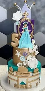 Three-tier Princess Jasmine themed cake in teal, gold, and purple with a gold number 5 and a white crown topper. Three-tier Princess Jasmine themed cake in teal, gold, and purple with a gold number 5 and a white crown topper.