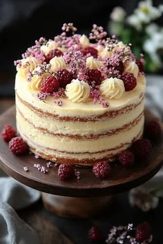 Naked cake with white soft icing, topped with fresh raspberries, pink filler flowers, and piped cream rosettes on a wooden stand.