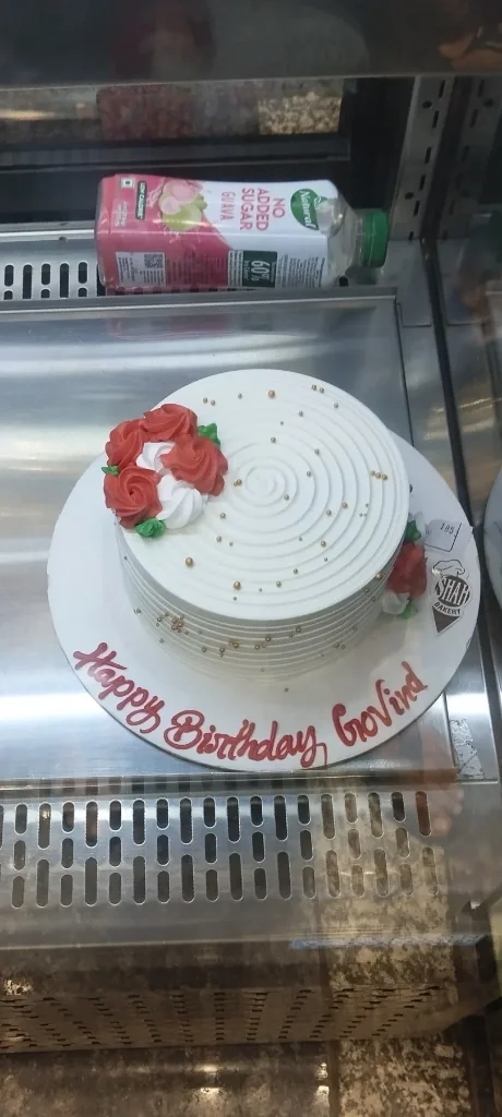 White round cake with red and white icing roses, gold dragees, and a Happy Birthday Govind message on the base board. White round cake with red and white icing roses, gold dragees, and a Happy Birthday Govind message on the base board.