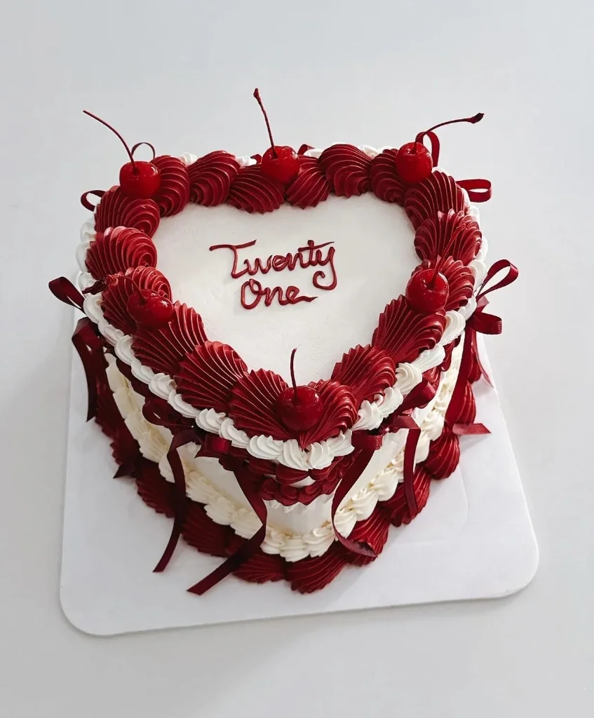 Heart-shaped white vintage cake with deep red piped icing ruffles, cherries, and red satin ribbons.