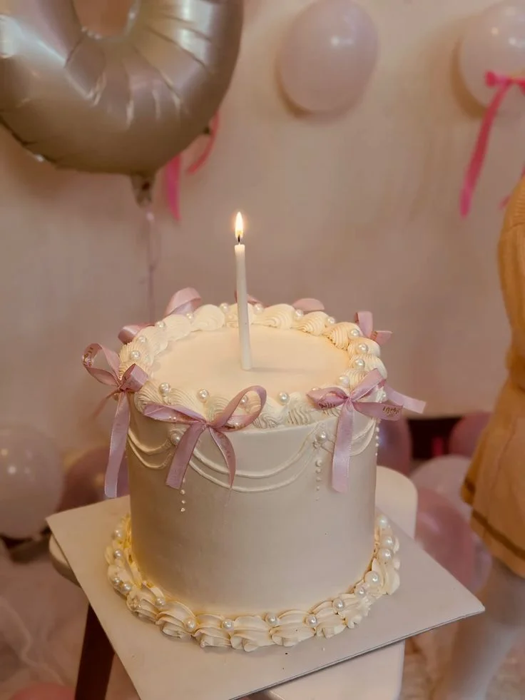 Elegant cream-colored tall cake with pink satin ribbons, white pearls, and a single lit taper candle on top. Elegant cream-colored tall cake with pink satin ribbons, white pearls, and a single lit taper candle on top.