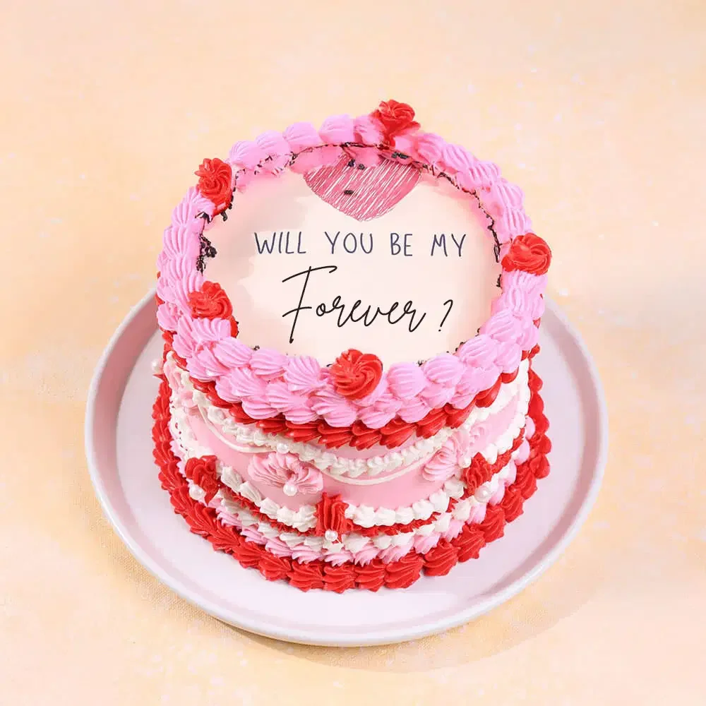 Pink and red vintage-style heart cake with ruffled icing borders and a 'Will You Be My Forever?' message on top.