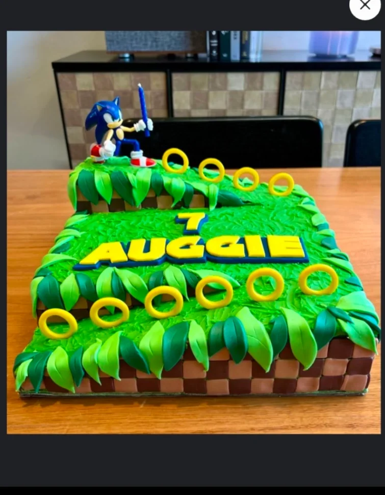 Sonic the Hedgehog themed square cake with green grass icing, checkered sides, golden rings, and a Sonic figurine holding a blue candle. Sonic the Hedgehog themed square cake with green grass icing, checkered sides, golden rings, and a Sonic figurine holding a blue candle.