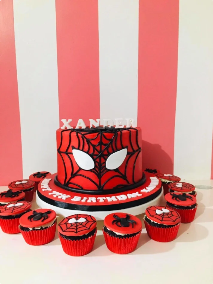 Red single tier Spiderman cake with black web icing and white eyes on a base board with cupcakes