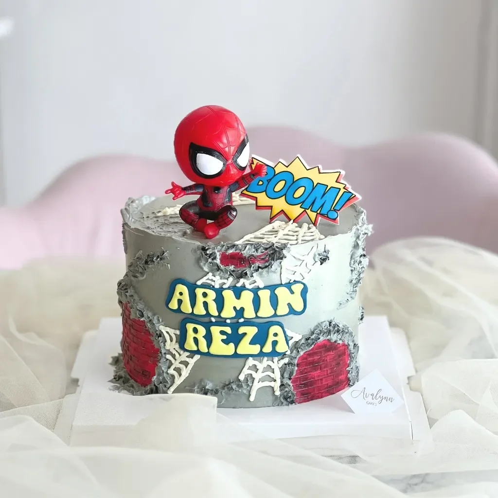 Spiderman-Inspired Silver Birthday Cake Silver 1-tier soft icing birthday cake with a Spiderman toy topper, BOOM sign, brick panels, and name lettering