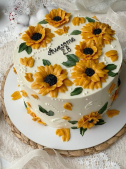 White round cake decorated with vibrant yellow icing sunflowers and green leaves, featuring a black icing message 'Awesome 26'. White round cake decorated with vibrant yellow icing sunflowers and green leaves, featuring a black icing message 'Awesome 26'.