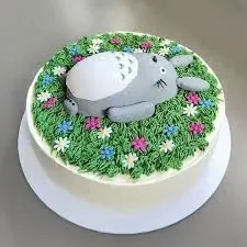 White round cake featuring a 3D fondant Totoro figure lying on a bed of green piped icing grass with small colorful flowers. White round cake featuring a 3D fondant Totoro figure lying on a bed of green piped icing grass with small colorful flowers.