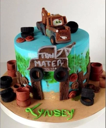 Disney Cars Tow Mater themed birthday cake with 3D fondant truck, tires, and wooden fence decorations on a blue sky background.