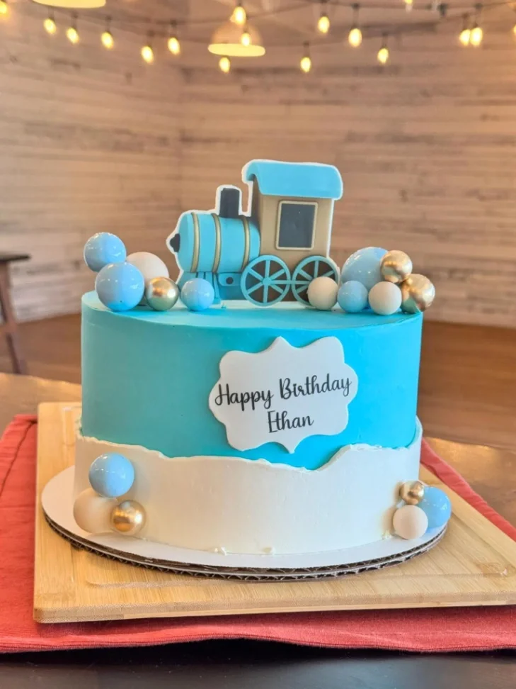 Light blue and white train themed birthday cake with a blue steam engine topper and gold and blue decorative spheres. Light blue and white train themed birthday cake with a blue steam engine topper and gold and blue decorative spheres.