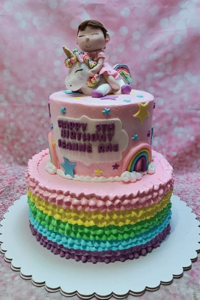 Two-tier pink birthday cake with a rainbow ruffle base, a fondant girl riding a unicorn topper, and rainbow and star decorations. Two-tier pink birthday cake with a rainbow ruffle base, a fondant girl riding a unicorn topper, and rainbow and star decorations.