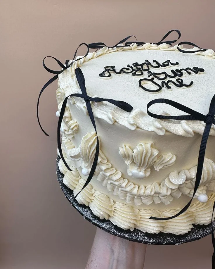 Elegant cream-colored vintage-style cake with intricate piped ruffles, black satin bows, and black icing calligraphy reading 'Regina turns One'.