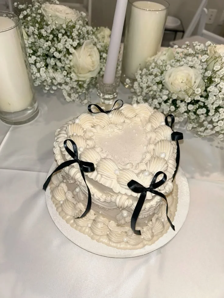 Heart-shaped vintage white cake with intricate buttercream piping, white pearls, and black satin bows on a white base.
