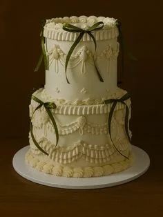 Elegant 2-tier ivory vintage-style cake with intricate piped ruffles, swags, and dark green satin ribbons.