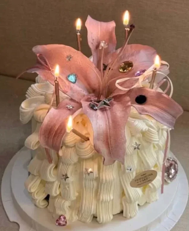 Vintage Lily White Floral Cake White 1-tier cake with soft icing, elaborate vertical piped white buttercream ruffles, a large pink lily flower topper, and tall thin candles