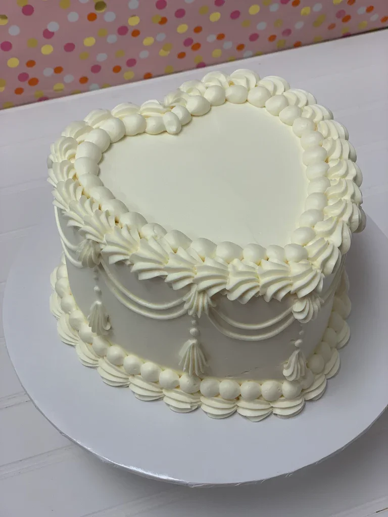 White heart-shaped vintage cake with intricate buttercream piping, ruffles, and tassels on a white base.