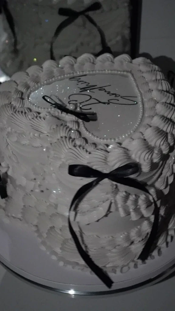 White heart-shaped vintage cake with intricate piped ruffles, black satin bows, and a personalized message on top. White heart-shaped vintage cake with intricate piped ruffles, black satin bows, and a personalized message on top.