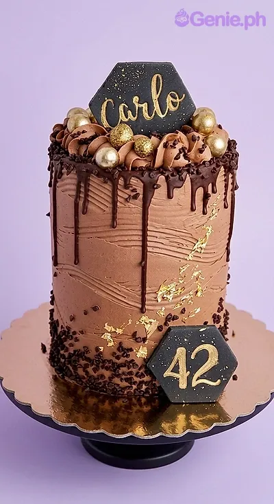 A brown 1-tier 42nd Birthday cake, with black hexagonal name and number toppers, chocolate drip, and gold leaf. A brown 1-tier 42nd Birthday cake, with black hexagonal name and number toppers, chocolate drip, and gold leaf.