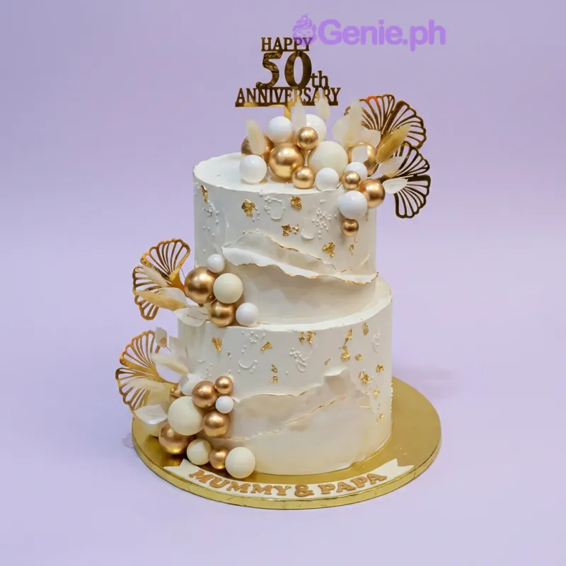 A white 2-tier 50th Anniversary cake, with a gold 50th topper, gold and white spheres, and gold leaves. A white 2-tier 50th Anniversary cake, with a gold 50th topper, gold and white spheres, and gold leaves.