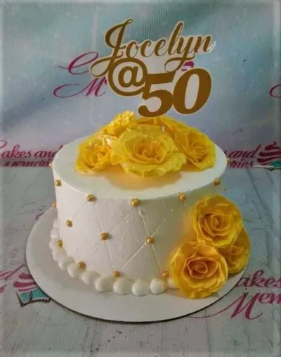 A white 1-tier 50th birthday cake, featuring a gold Jocelyn @ 50 topper, yellow roses, and a quilted pattern. A white 1-tier 50th birthday cake, featuring a gold Jocelyn @ 50 topper, yellow roses, and a quilted pattern.