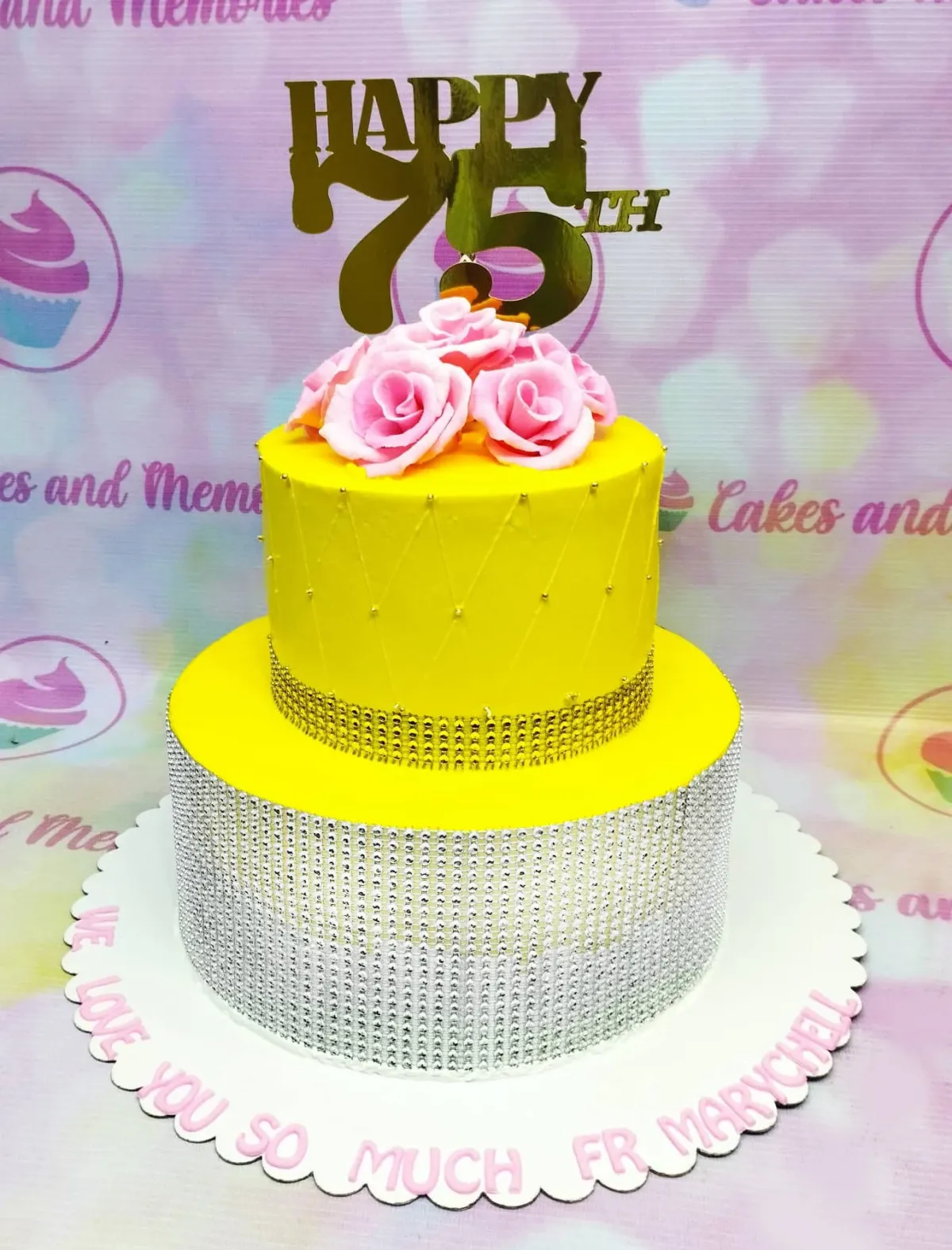 A yellow and pink 2-tier cake themed for 75th Birthday, featuring a gold 'Happy 75th' topper, pink roses, and a silver wrap. A yellow and pink 2-tier cake themed for 75th Birthday, featuring a gold 'Happy 75th' topper, pink roses, and a silver wrap.