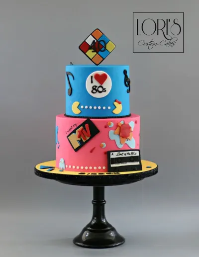 Two-tier 80s themed fondant cake with blue and pink tiers, featuring a Rubik's Cube topper, MTV logo, and Pac-Man decorations. Two-tier 80s themed fondant cake with blue and pink tiers, featuring a Rubik's Cube topper, MTV logo, and Pac-Man decorations.