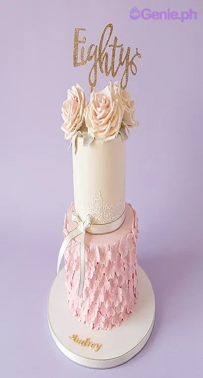 A pink and ivory 2-tier fondant cake for an 80th birthday, with a gold 'Eighty' topper, peach roses, lace, and pink petal details. A pink and ivory 2-tier fondant cake for an 80th birthday, with a gold 'Eighty' topper, peach roses, lace, and pink petal details.