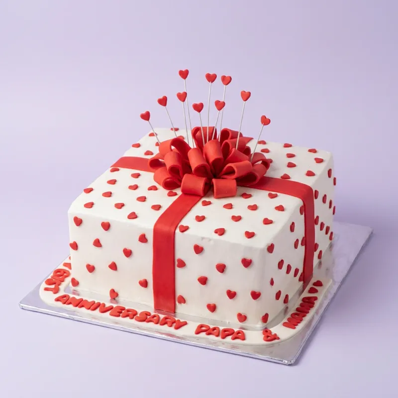 A white and red square cake themed as an anniversary gift box, featuring a large red fondant bow and small red hearts. A white and red square cake themed as an anniversary gift box, featuring a large red fondant bow and small red hearts.