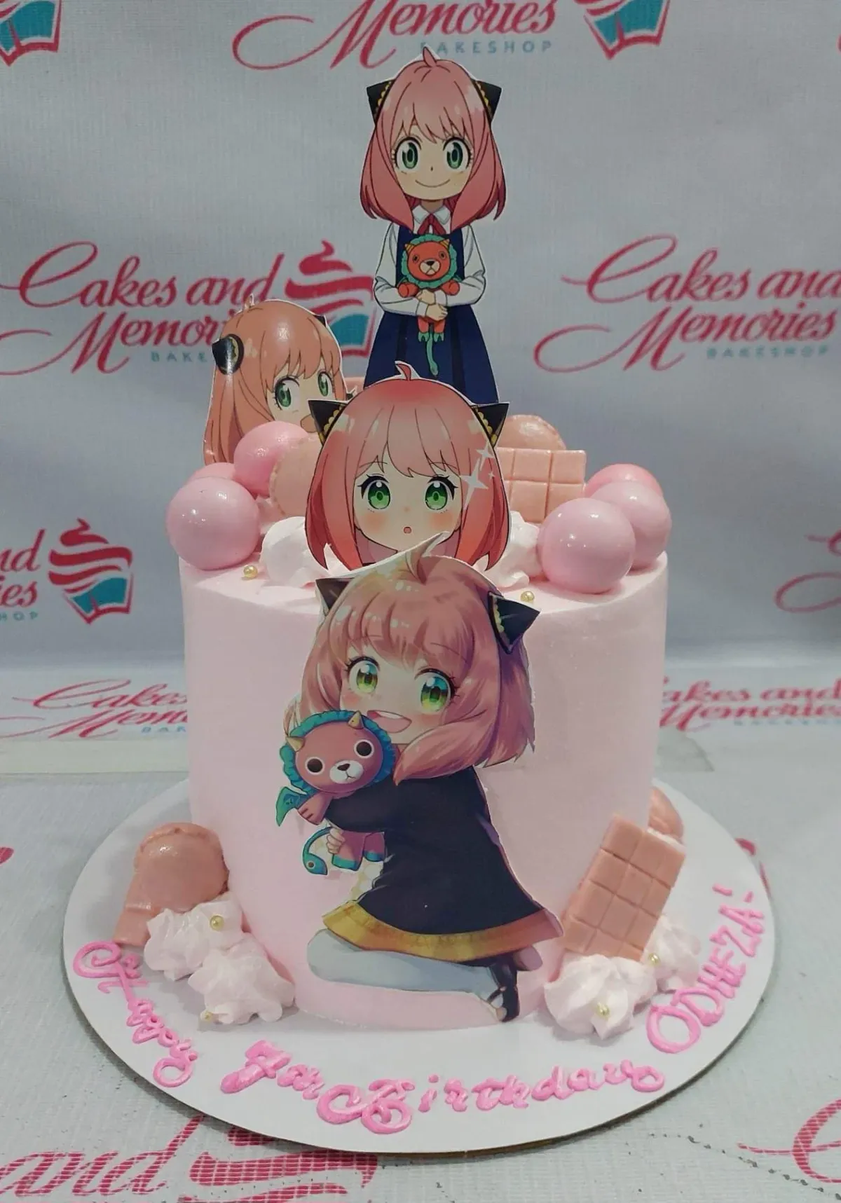 A pink and white 1-tier cake themed around Anya Forger, featuring character printouts, pink chocolate bars, and macarons. A pink and white 1-tier cake themed around Anya Forger, featuring character printouts, pink chocolate bars, and macarons.