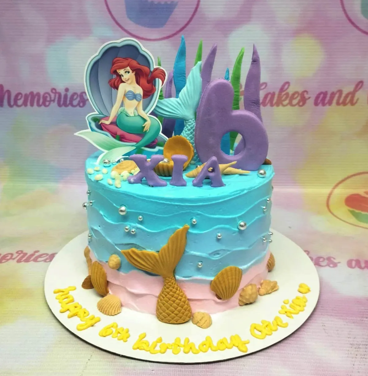A blue and pink 1-Tier cake themed Ariel Mermaid, featuring Ariel, a purple '6', gold mermaid tail, and edible seashells. A blue and pink 1-Tier cake themed Ariel Mermaid, featuring Ariel, a purple '6', gold mermaid tail, and edible seashells.