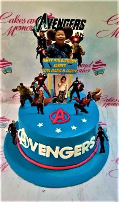 A blue 1 tier cake featuring Avengers character graphics, a child's photo, and a birthday banner, decorated with a red 'A' logo and white stars. A blue 1 tier cake featuring Avengers character graphics, a child's photo, and a birthday banner, decorated with a red 'A' logo and white stars.