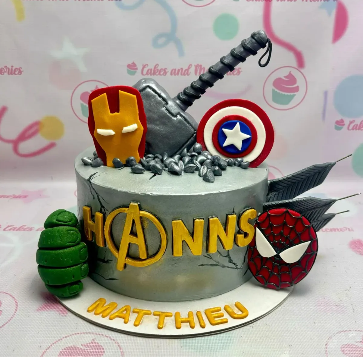 A gray 1 Tier Fondant Avengers cake, featuring Thor's hammer, Iron Man mask, and Captain America shield. A gray 1 Tier Fondant Avengers cake, featuring Thor's hammer, Iron Man mask, and Captain America shield.