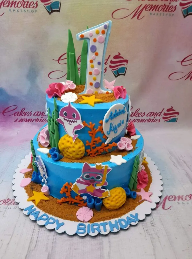 Two-tier blue Baby Shark themed birthday cake with edible underwater decorations, character printouts, and a large number one topper. Two-tier blue Baby Shark themed birthday cake with edible underwater decorations, character printouts, and a large number one topper.