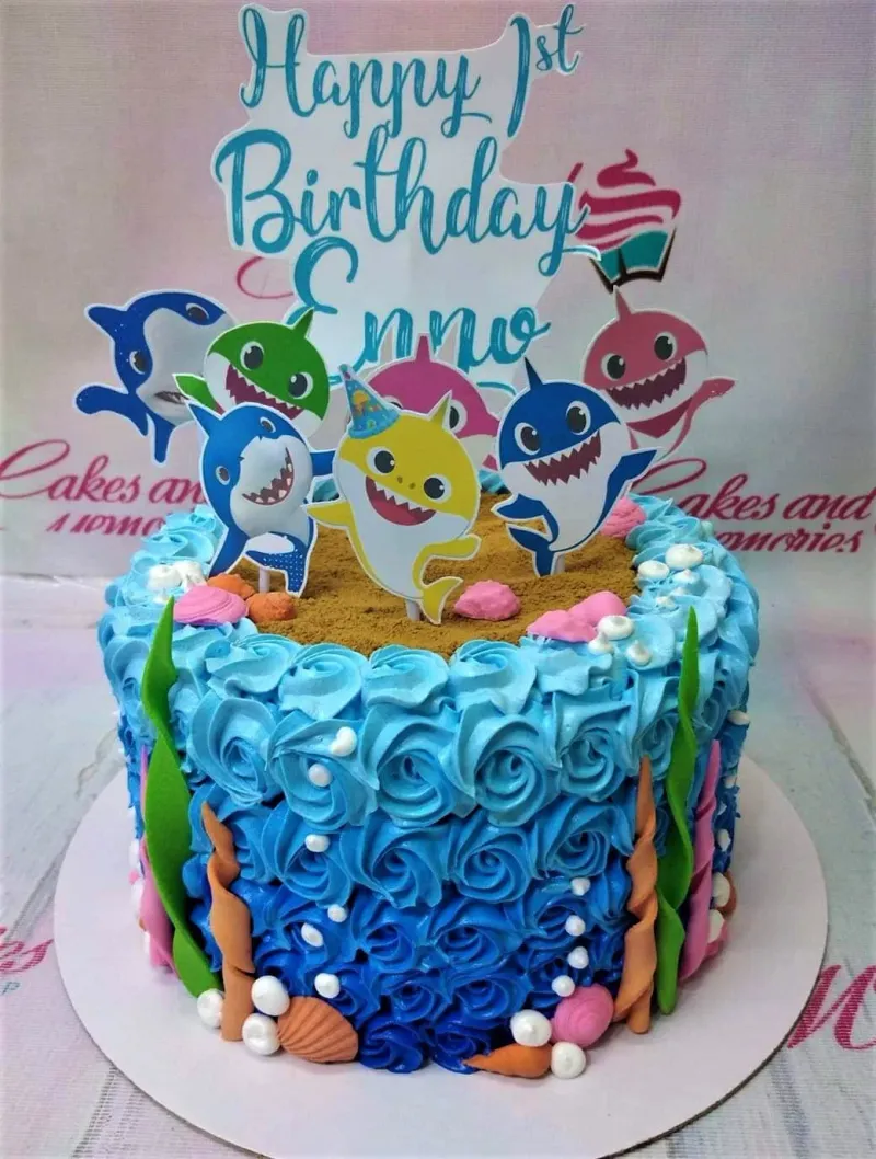 Baby Shark themed birthday cake with blue icing rosettes, sand-textured top, and colorful shark character printouts. Baby Shark themed birthday cake with blue icing rosettes, sand-textured top, and colorful shark character printouts.