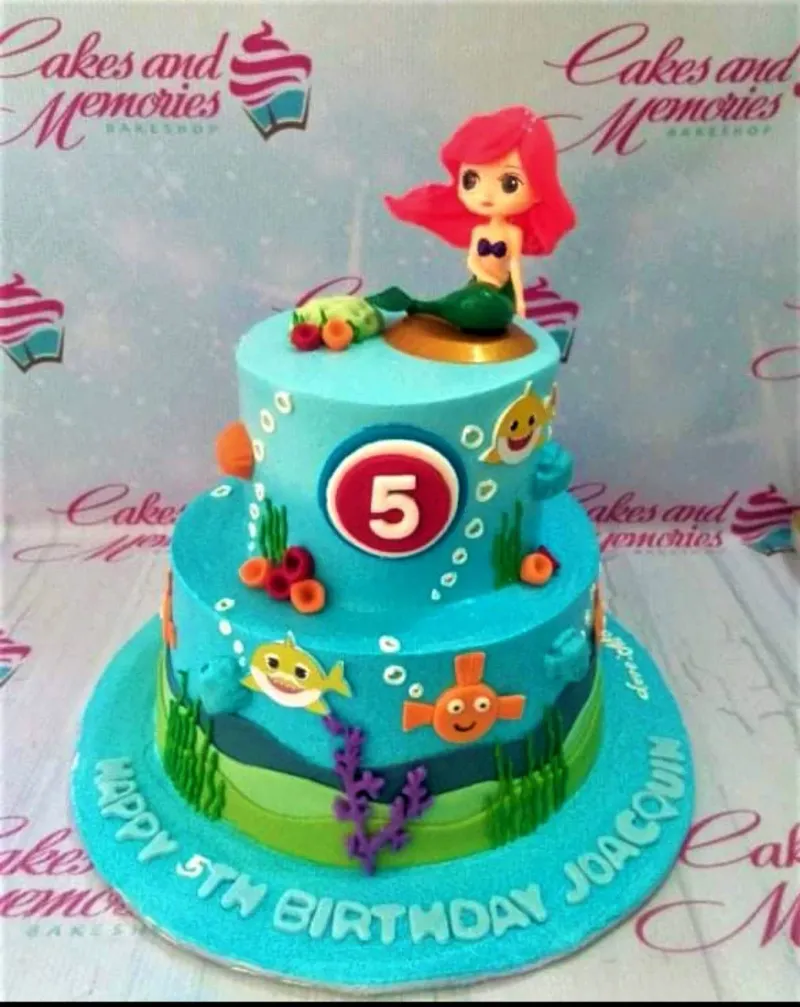 Two-tier blue Baby Shark and Mermaid cake with a mermaid toy, number 5, and fondant sea elements. Two-tier blue Baby Shark and Mermaid cake with a mermaid toy, number 5, and fondant sea elements.