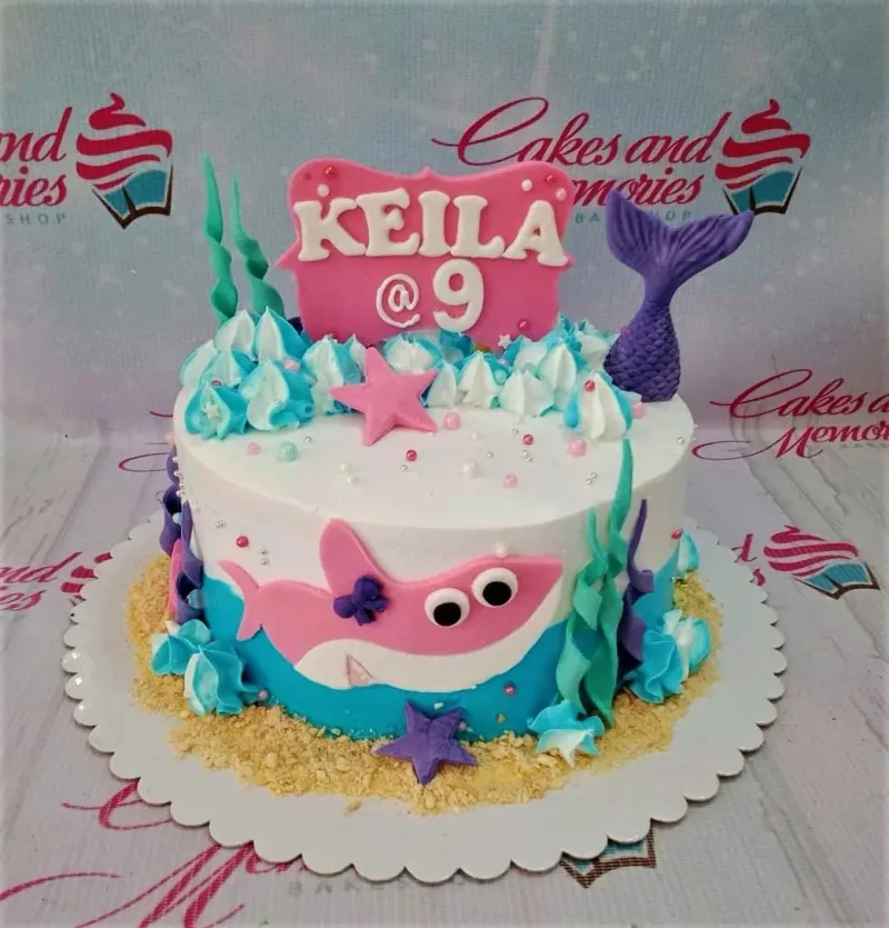 A white and blue 1-tier cake themed Baby Shark Mermaid, featuring a pink shark, purple mermaid tail, seaweed, and starfish. A white and blue 1-tier cake themed Baby Shark Mermaid, featuring a pink shark, purple mermaid tail, seaweed, and starfish.