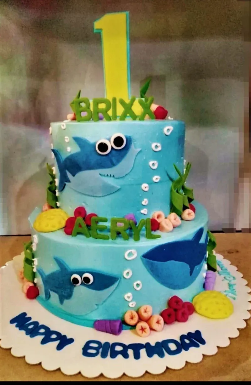 Two-tier blue Baby Shark themed birthday cake with fondant sharks, coral, and seaweed decorations. Two-tier blue Baby Shark themed birthday cake with fondant sharks, coral, and seaweed decorations.