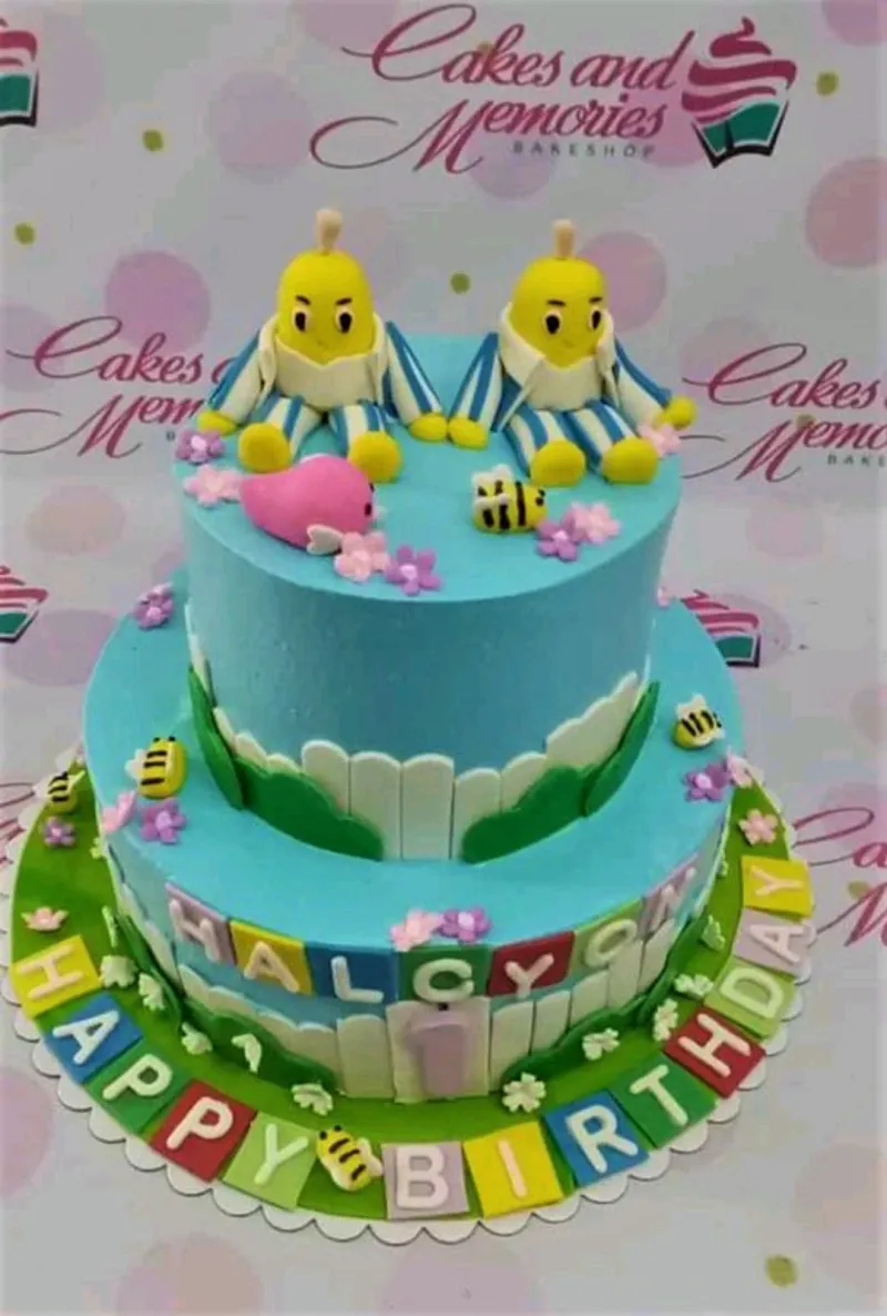 Two-tier blue Bananas in Pyjamas themed birthday cake with fondant banana characters, bees, and a pink whale. Two-tier blue Bananas in Pyjamas themed birthday cake with fondant banana characters, bees, and a pink whale.