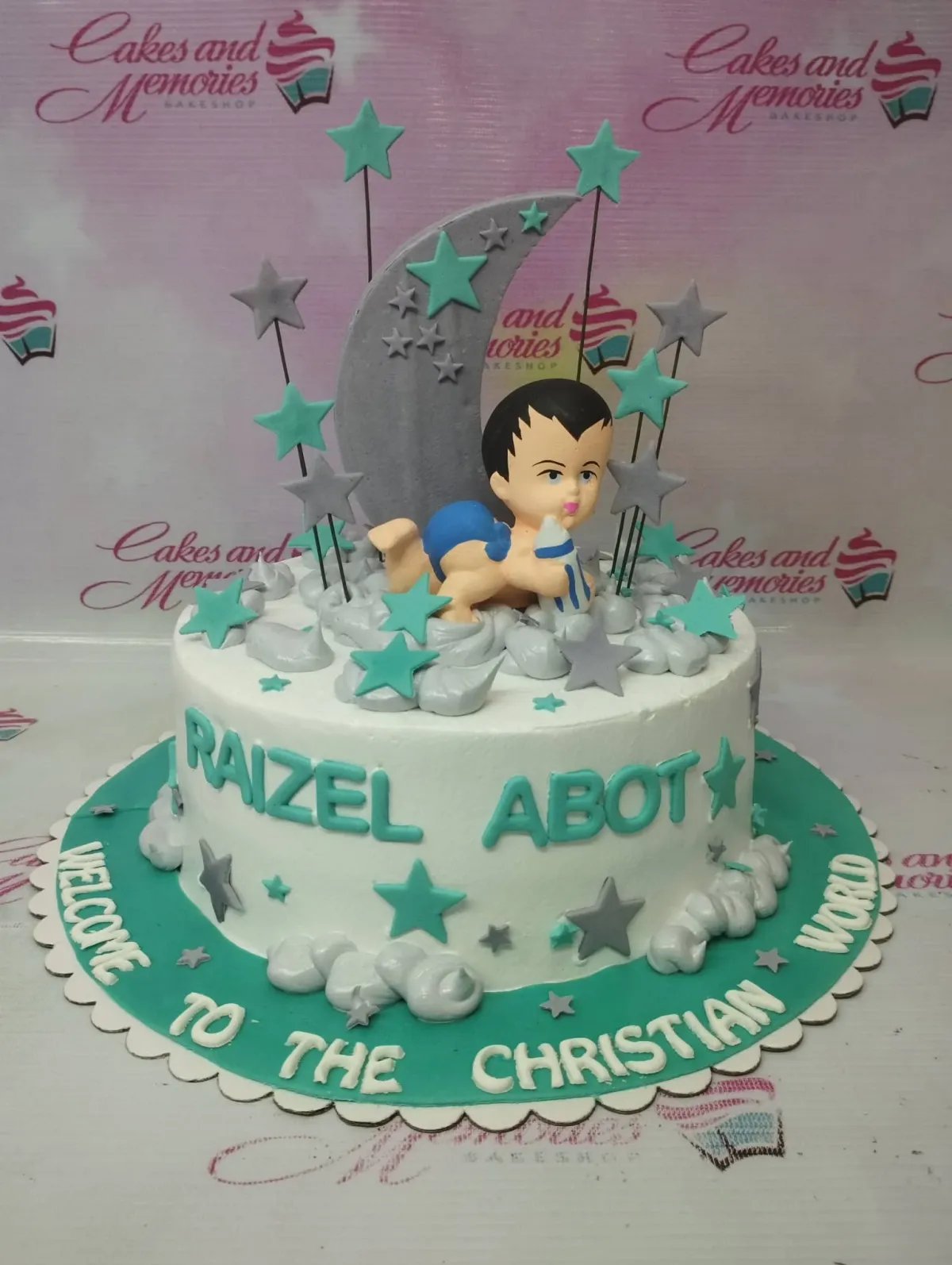 White 1-tier baptismal cake with a 3D fondant baby figure, moon, and teal and gray stars on a teal base board. White 1-tier baptismal cake with a 3D fondant baby figure, moon, and teal and gray stars on a teal base board.